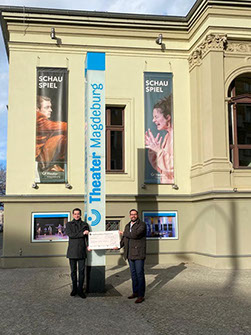 Zwei Personen mit einem Spendenscheck vor dem Schauspielhaus des Theaters Magdeburg