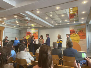 Podiumsrunde vor Publikum in einem Tagungsraum, im Hintergrund Plakate „Engagiert in und für Ostdeutschland"